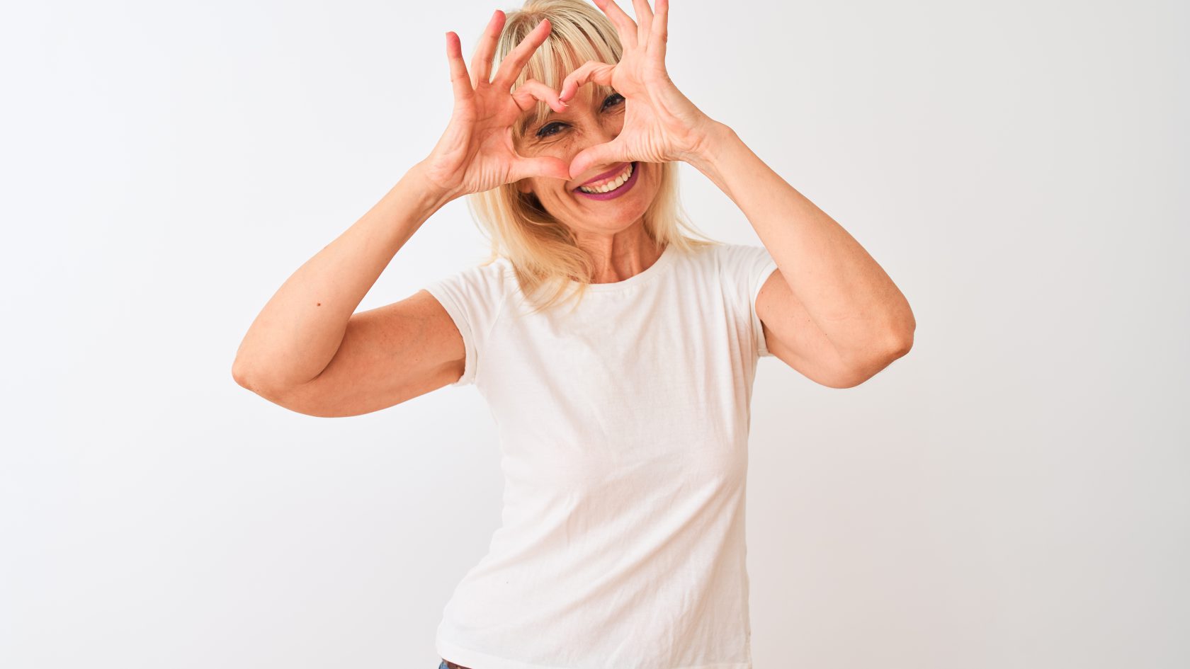 Middle age woman wearing casual t-shirt standing over isolated white background Doing heart shape with hand and fingers smiling looking through sign